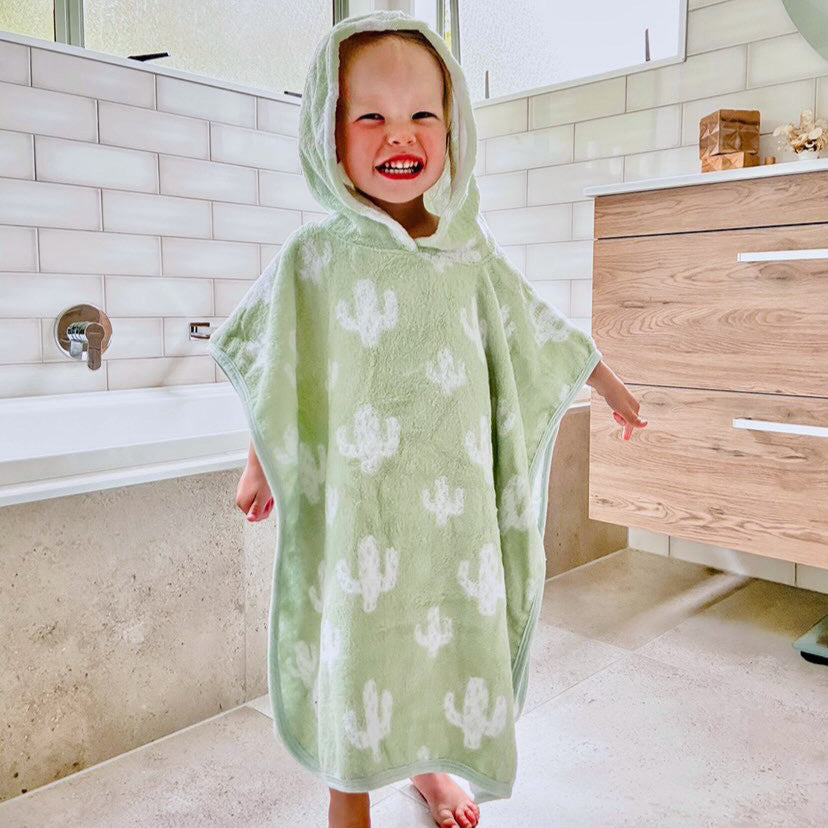 A child wearing a green hooded poncho with white cacti pattern, standing in a bathroom setting.
