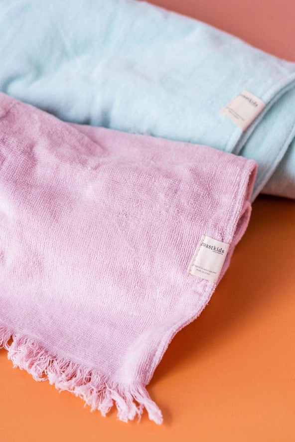 Two folded towels, one pink and one light blue, on an orange surface with a visible brand label.