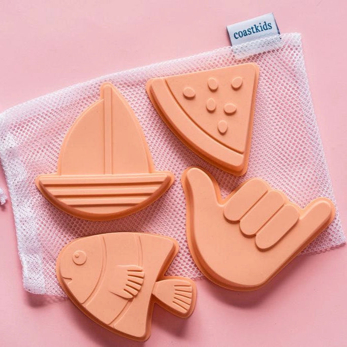 Set of silicone sand toys shaped like a sailboat, fish, watermelon slice, and hand in a mesh storage bag on a pink background.
