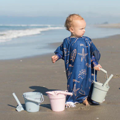 Child in a blue dress with starfish patterns playing on a sandy beach with buckets.