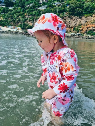 Child in a floral outfit and hat standing in shallow water near a coastal area.