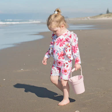 Child in a floral swimsuit on a beach holding a pink bucket