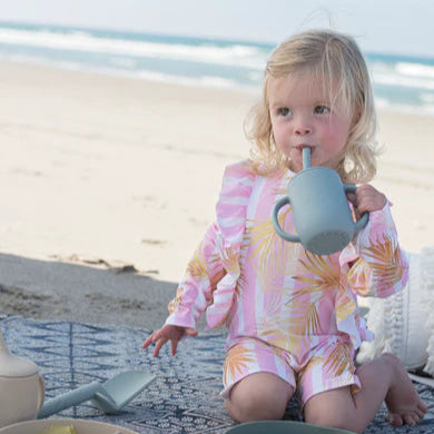 Child in a colorful swimsuit sitting on a beach with a teapot and food containers.