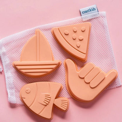 Set of silicone sand toys shaped like a sailboat, fish, watermelon slice, and hand in a mesh storage bag on a pink background.