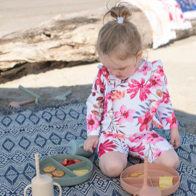 Child in a floral swimsuit sitting on a patterned blanket with picnic items at the beach.